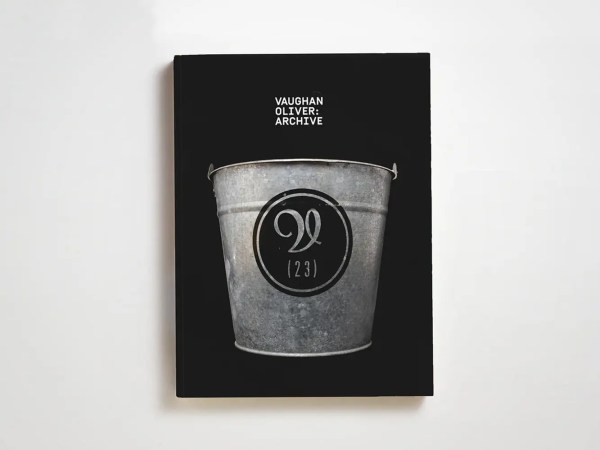 A book with a black cover featuring a large image of a metal bucket. Above the bucket, the text reads VAUGHAN OLIVER: ARCHIVE. Inside the bucket is a black circle with a stylized “V” and “(23)” beneath it.