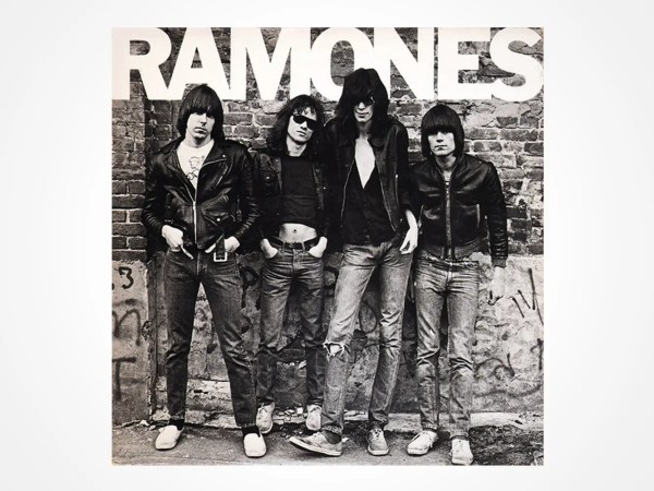 Four men in leather jackets and jeans stand against a graffiti-covered brick wall beneath the large text RAMONES. The image is black and white, and the group appears relaxed with a punk rock style.