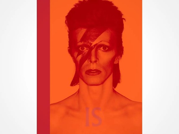 A stylized, red-and-orange-tinted image of a person with a mullet hairstyle, lightning bolt face paint, and bare shoulders. The word IS appears at the bottom of the image.