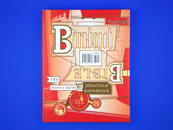 The signed hardback book "Jonathan Barnbrook – The Barnbrook Bible" features bold, layered text and abstract design elements by Jonathan Barnbrook on a red and beige cover, set against a blue background.