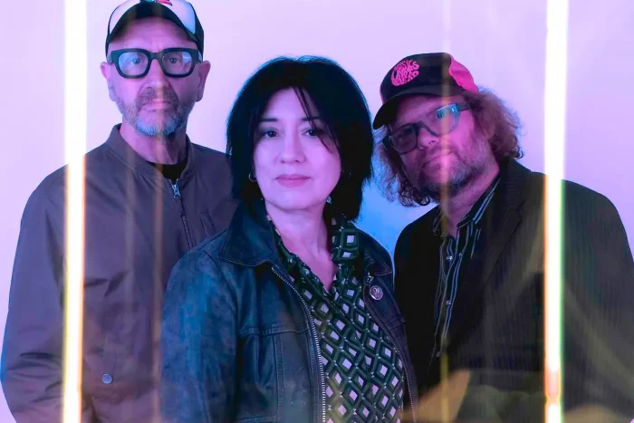 Three people stand together against a light purple background, embodying the spirit of a Home Template. Two men wear glasses, jackets, and hats; a woman with dark hair stands center in a patterned shirt and jacket. Vertical light streaks cross the image.