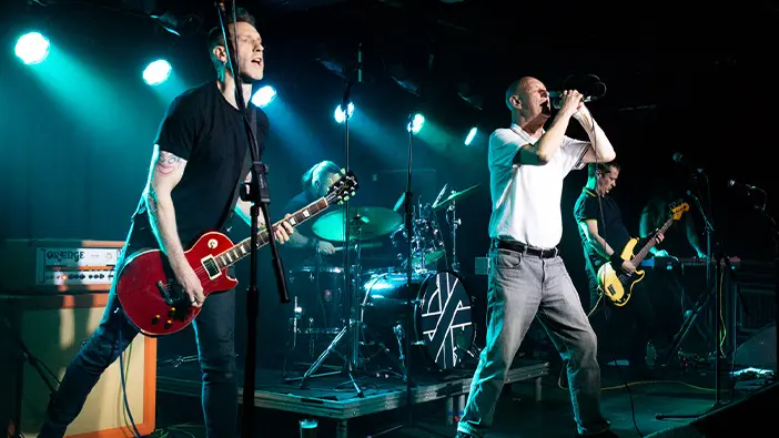 A rock band performs on stage under blue lights, creating an electrifying atmosphere. The guitarist plays a red electric guitar while the drummer, vocalist, and bassist bring the performance to life.